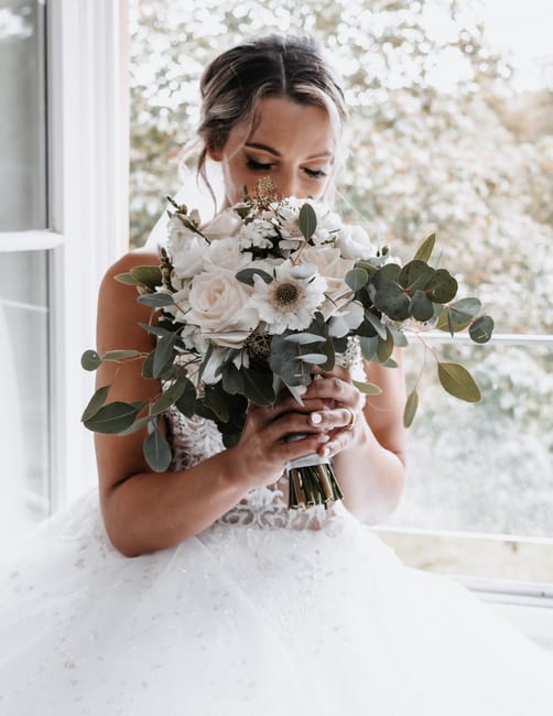 A bride holding a large, elegant bouquet featuring white roses, anemones, and eucalyptus leaves. The bouquet is wrapped in a metallic gold ribbon, and the flowers appear fresh with delicate petals and