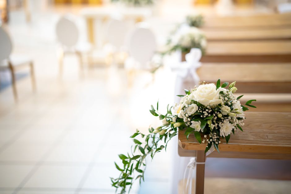 A close-up of a wedding floral arrangement resting on a wooden church pew, featuring a bouquet of fresh white roses, lisianthus, and baby's breath with lush green foliage, tied with a white ribbon. Th