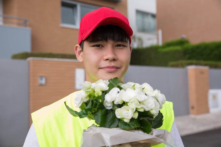 A young male delivery person with light skin, wearing a bright yellow high-visibility vest and a red baseball cap, stands outdoors in front of modern residential buildings with brick and grey facades.