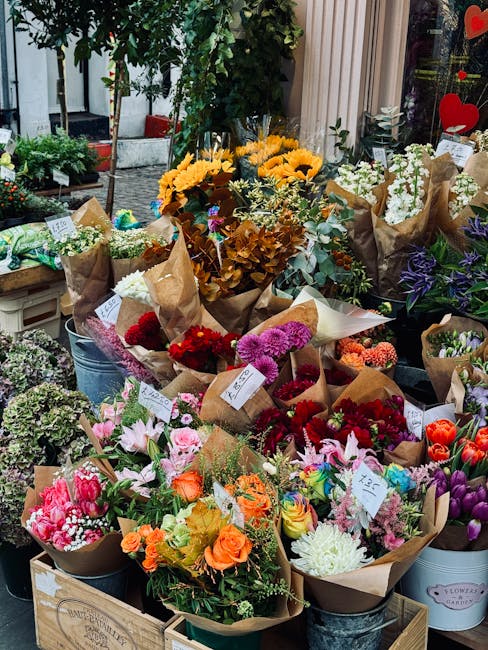 An outdoor floral display featuring a variety of fresh flower bouquets arranged on wooden crates and metal buckets. The bouquets showcase vibrant colours including bright yellow sunflowers, deep red r