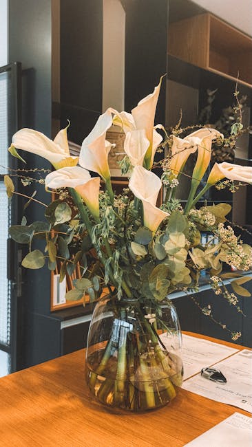 A large, elegant floral arrangement in a clear glass vase featuring pale peach calla lilies with smooth, curved petals and fresh green stems partially submerged in water. The bouquet includes sprigs o