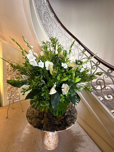 A lush floral arrangement featuring predominantly white flowers, including lilies and anemones, complemented by various shades of green foliage with broad and slender leaves. The bouquet is set in a d